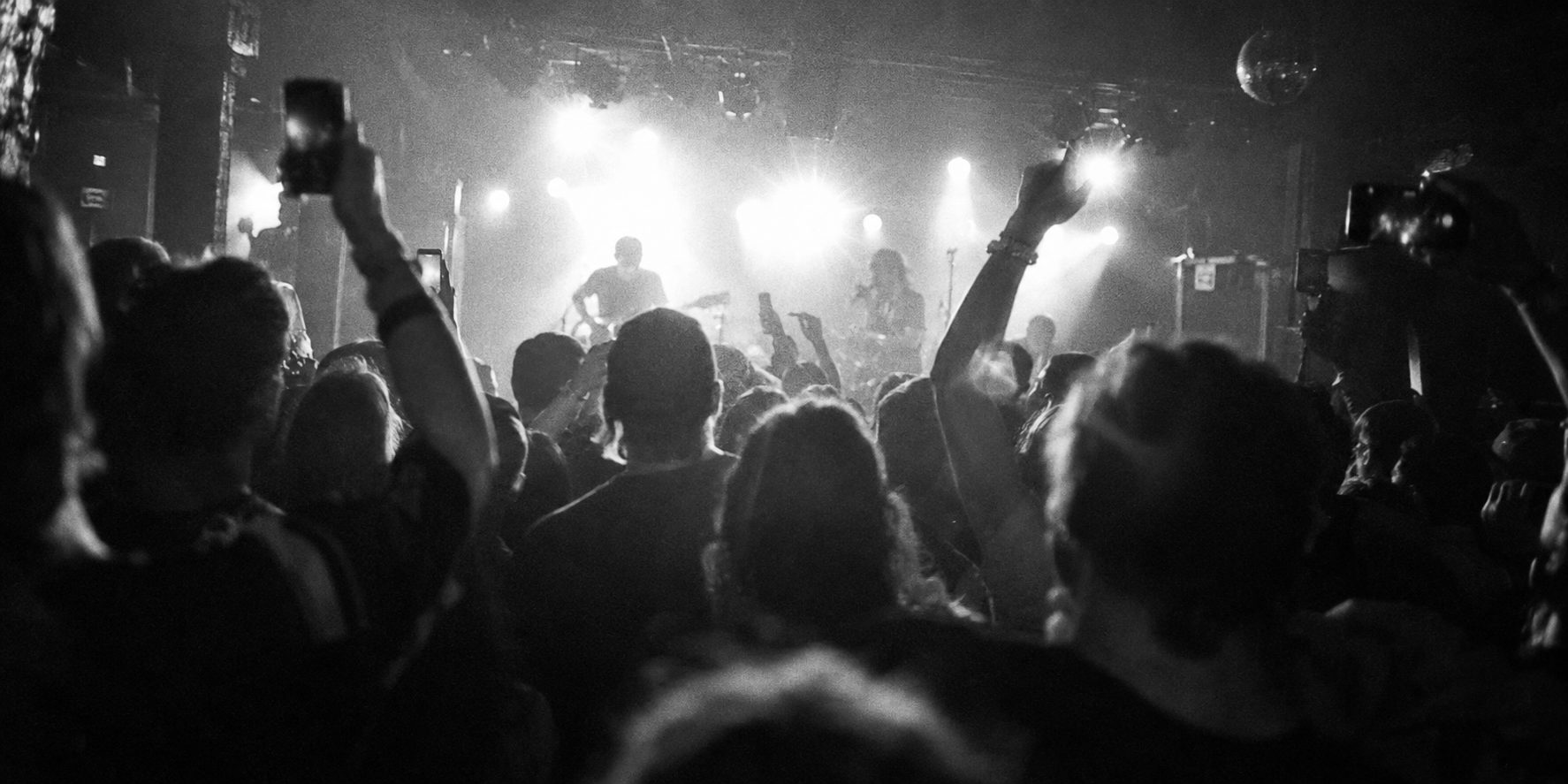 A crowd at a small venue seen from behind, phones raised, stage light flaring
