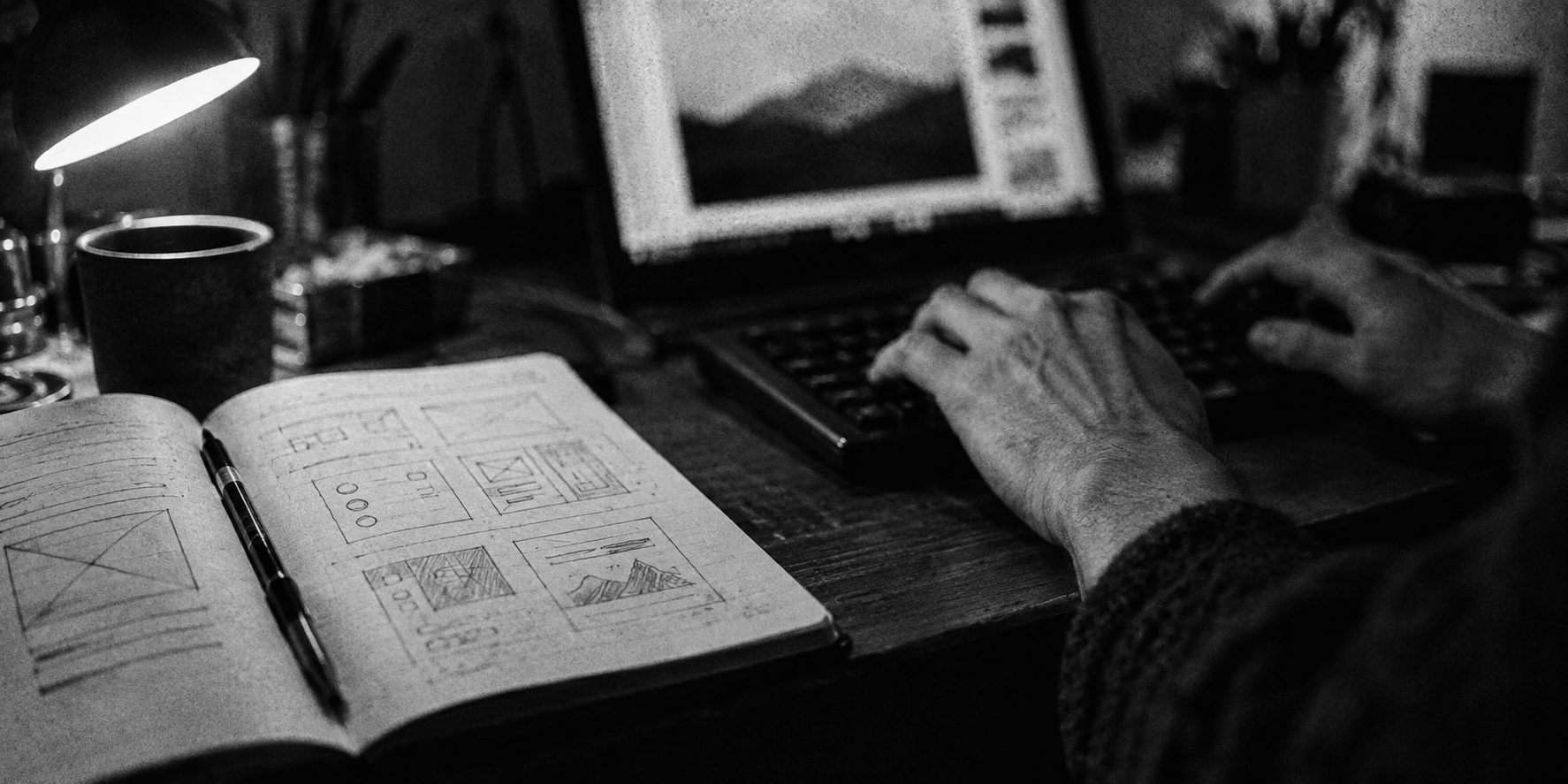 Hands at a keyboard beside a notebook of hand-drawn wireframes under a desk lamp